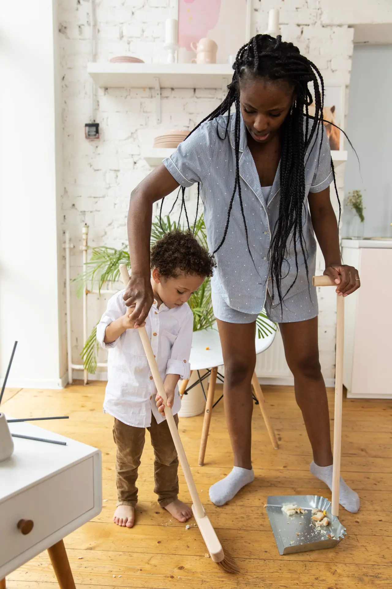 A mother and her young child sweeping the wooden floor of their living room, engaging in family chores.